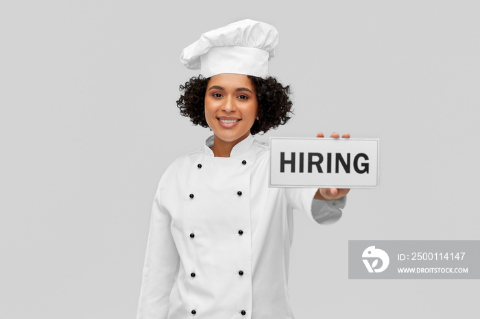 job, work and employment concept - happy smiling female chef in white toque and jacket holding hiring sign over grey background