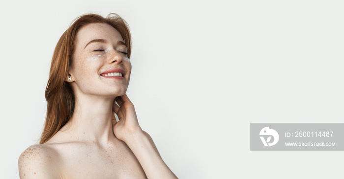 Ginger lady with freckles is wearing anti aging eye patches posing on white studio wall with naked shoulders advertising something