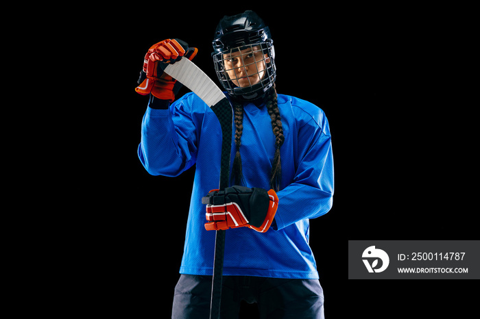 Young female hockey player isolated on black background. Sportswoman wearing equipment and helmet standing with the stick. Concept of sport, healthy lifestyle, motion, movement, action.