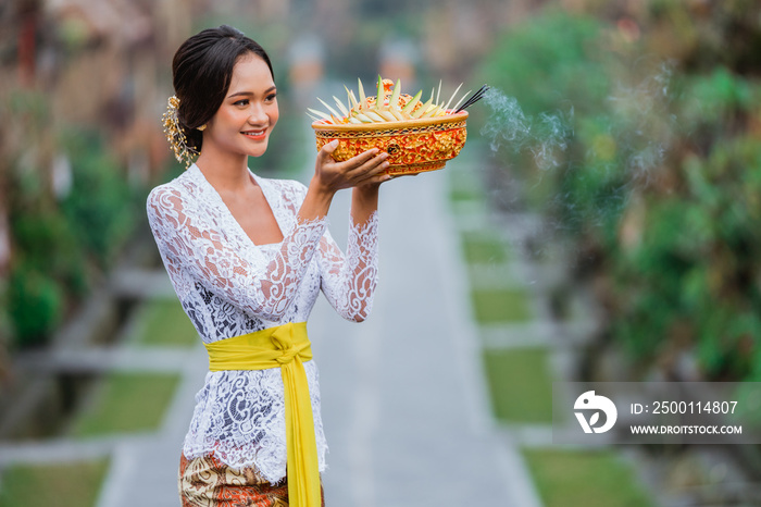 beautiful balinese woman wearing traditional kebaya in the penglipuran bali village smiling while carrying offering for praying