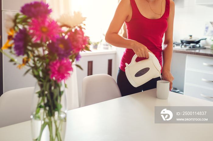 Woman pouring hot water from electric kettle in kitchen. Girl making tea. Modern kitchen design.