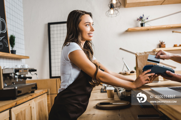 attractive cashier standing in brown apron and holding terminal wile man paying with credit card in coffee house