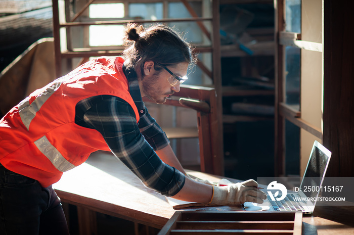 Young and handsome caucasian carpenter in the carpentry shop is working and checking blueprint in laptop to repair a furniture.