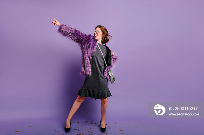 Full-length portrait of enchanting glamorous woman dancing in purple winter jacket. Studio shot of relaxing young lady with curly hairstyle posing on colorful background.