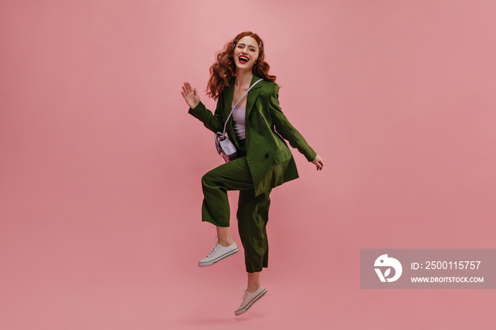 Cheerful caucasian young woman moves energetically while in photo studio with place for text. Red-haired curly beauty bounces in trendy green suit with small bag hanging over shoulder.