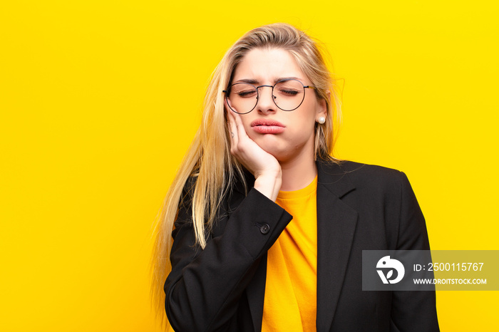 young pretty blonde woman holding cheek and suffering painful toothache, feeling ill, miserable and unhappy, looking for a dentist against yellow wall