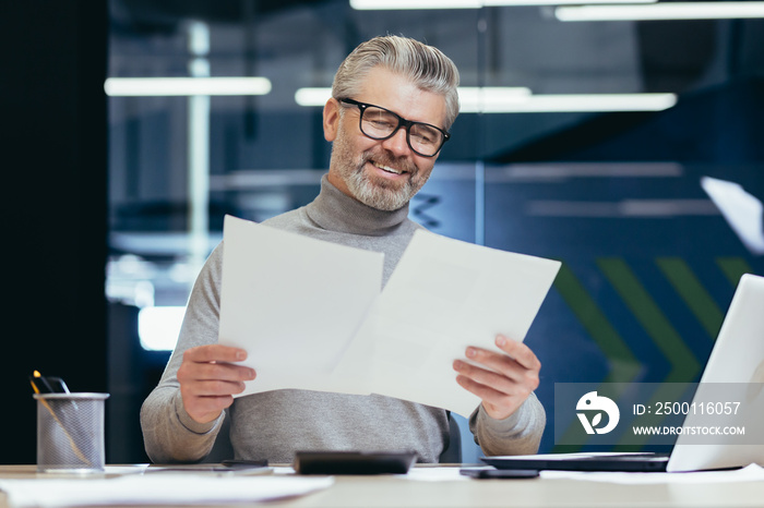 Senior smiling gray-haired businessman working in office at desk with laptop and documents. Holds in his hands, looks at sheets of paper, agreements, bills.