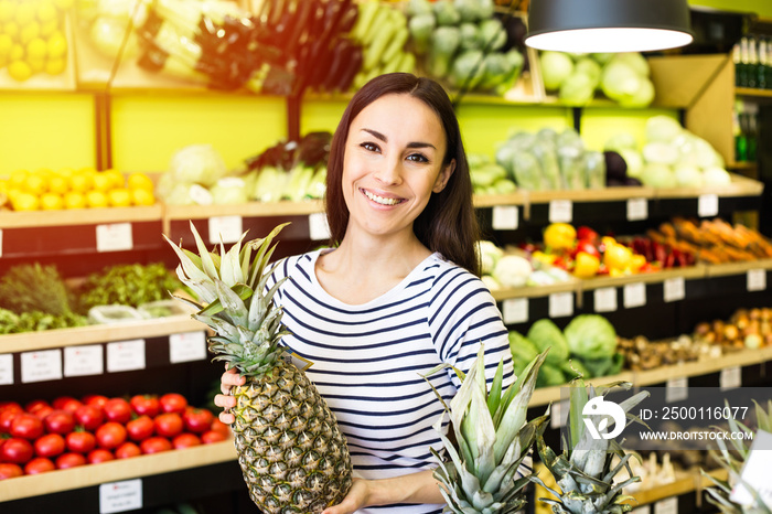 Attractive smiling young girl in casual clothes selects fruits and vegetables from a shelf in a grocery store.