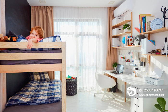 happy boy lies on the upper level of the bunk bed in bright kids room