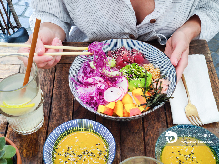 Woman eating tasty colorful healthy natural organic vegetarian Hawaiian poke bowl using asian chopsticks on rustic wooden table. Healthy natural organic eating concept.