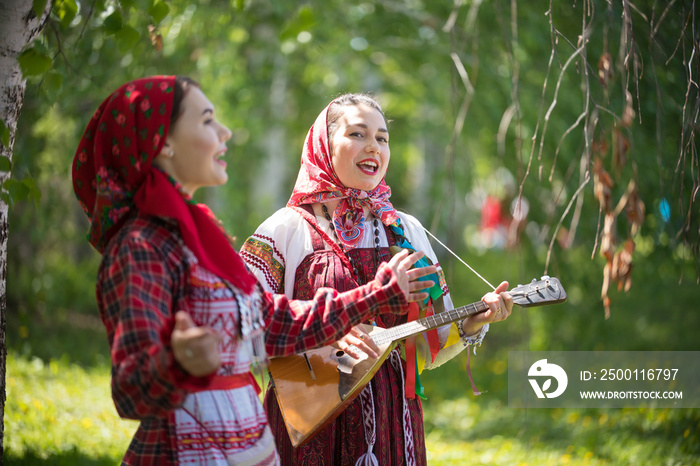 Two young woman in traditional russian clothes singing in the forest. One of them playing balalaika