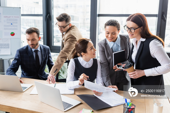 Smiling interracial businesswomen looking at document near businessmen using laptop