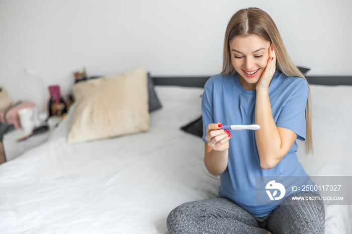 Smiling and excited girl checking her recent pregnancy test, sitting on bed couch at home, copy space