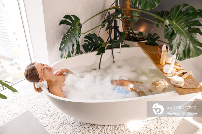 Relaxed young woman bathing with body care cosmetics on wooden shelf over modern white bubble filled bathtub at home. Spa, wellness concept