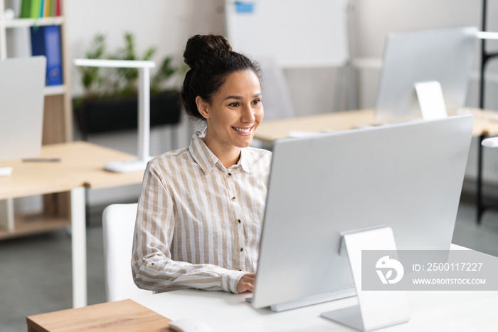 Happy latin businesswoman working with modern computer, checking email, sitting at her workplace in office interior