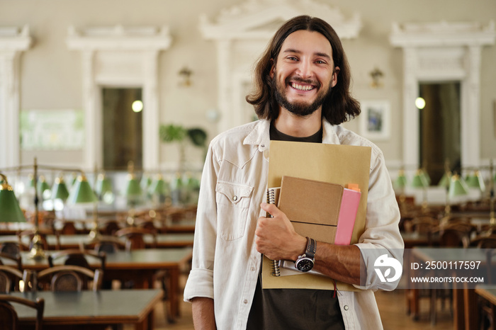 Portrait of young attractive male student with book joyfully looking in camera in library of university