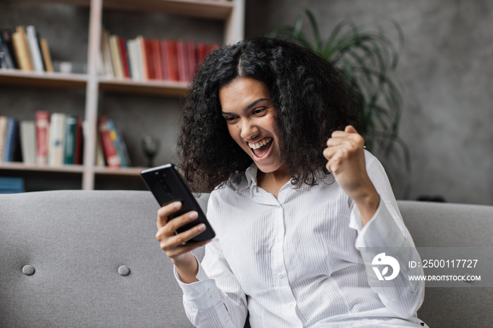 Attractive young woman in casual wear sitting on comfy couch with modern smartphone in hands. Smiling female person surfing internet during leisure time at home.