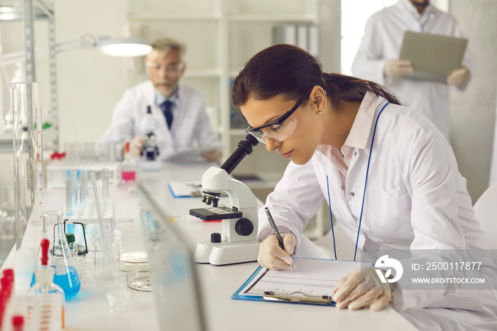 Serious scientist in lab coat, goggles and gloves doing advanced research, conducting laboratory experiment, looking in optical microscope, collecting data, taking notes on clipboard, writing report