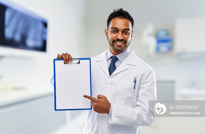 medicine, dentistry and profession concept - smiling indian male dentist in white coat with blank paper on clipboard over dental clinic office background