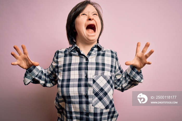 Young down syndrome woman wearing casual shirt over pink background crazy and mad shouting and yelling with aggressive expression and arms raised. Frustration concept.