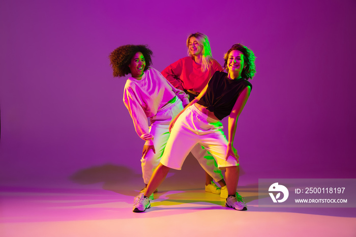 Flexible. Sportive girls dancing hip-hop in stylish clothes on purple-pink background at dance hall in green neon light. Youth culture, movement, style and fashion, action. Fashionable portrait.