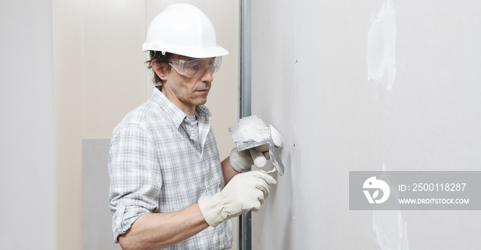 man drywall worker or plasterer putting plaster on plasterboard wall using a trowel and a spatula, fill the screw holes, wearing white hardhat, work gloves and safety glasses.