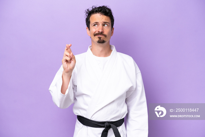 Young caucasian man doing karate isolated on purple background with fingers crossing and wishing the best