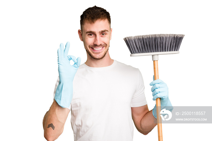 Young man holding a broom to clean his house cut out isolated cheerful and confident showing ok gesture.