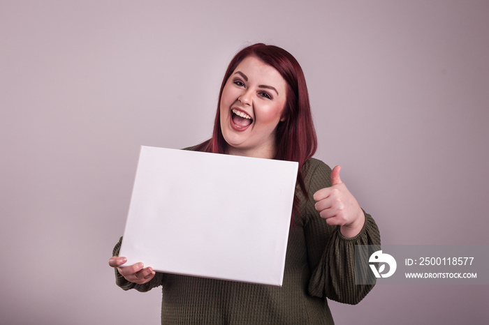 Young woman holding an empty white board giving a thumbs up excited facial  expression