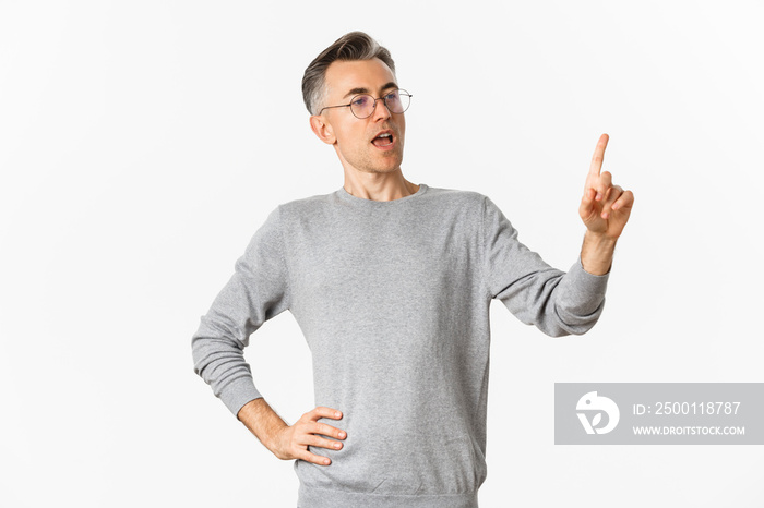 Image of confident middle-aged businessman in grey sweater and glasses, pointing finger and making speech, giving tasks to employees, standing over white background