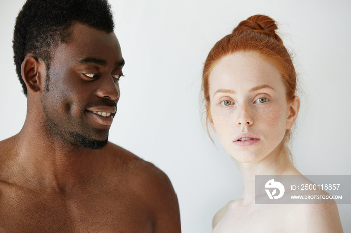 Portrait of happy loving interracial couple: shirtless African man looking at his nude redhead Caucasian girlfriend with love and affection, while she is standing next to him and looking at the camera