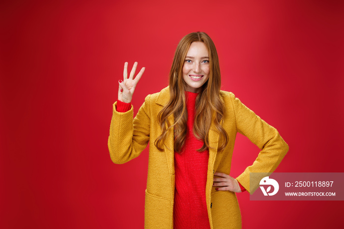 Cute friendly-looking ginger girl showing three reasons buy product smiling broadly making third reservation, looking delighted and confident in her choice posing against red background in yellow coat