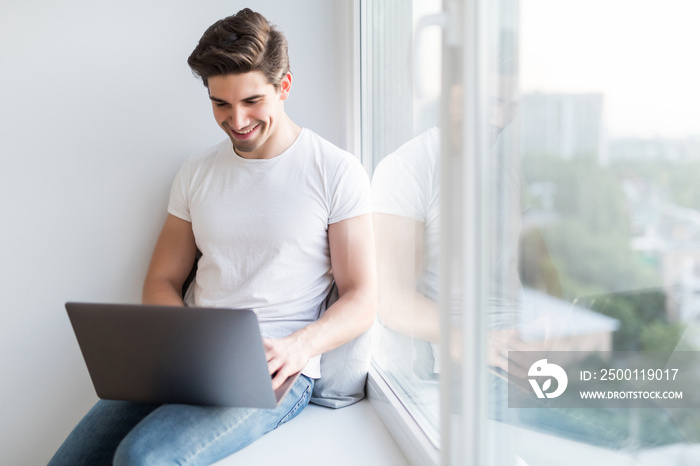 Young man working on laptop and sitting on windowsill