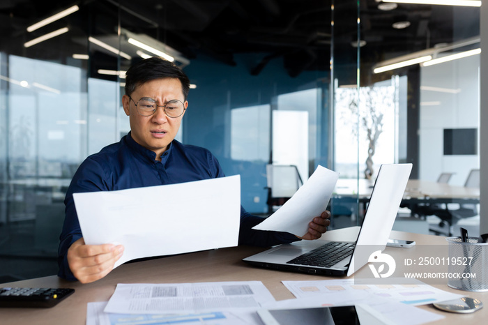 Asian businessman not satisfied with achievement results, boss looking at documents and upset, man in glasses and shirt working inside office with paperwork using laptop.