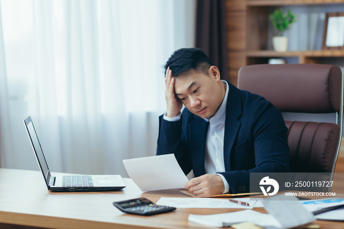 Young asian male accountant, office worker working with documents, tired, sitting at desk in office