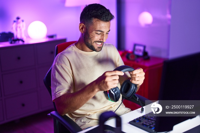 Young hispanic man streamer smiling confident holding headphones at gaming room