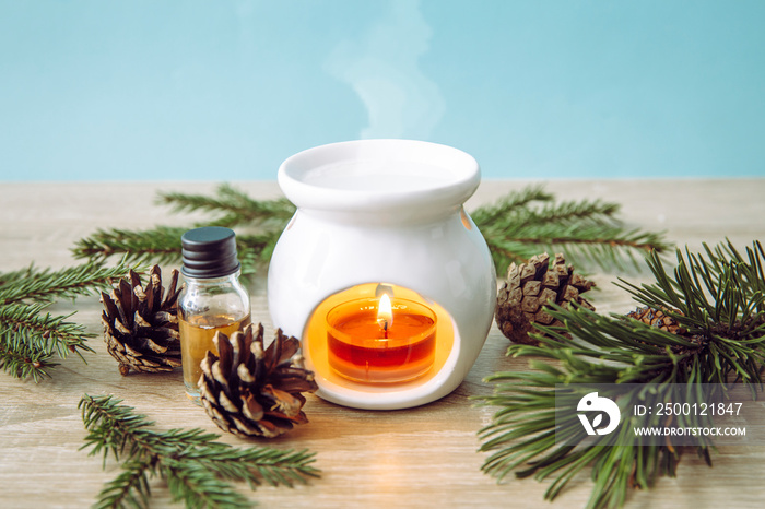 White ceramic candle aroma oil lamp with pine spruce tree essential oil bottle and tree branches for decoration on wooden and blue background.