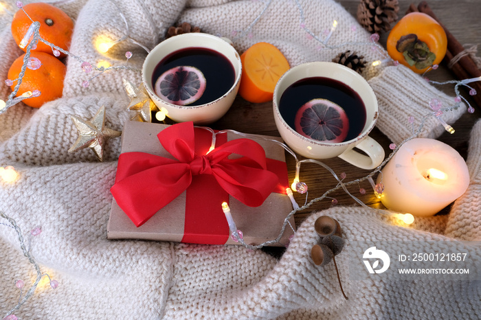 two cups of hot mulled wine with lemon, persimmon and tangerine fruits, candles, box with gift in kraft paper, with red bow lies on table, concept of holiday, congratulations merry christmas, new year