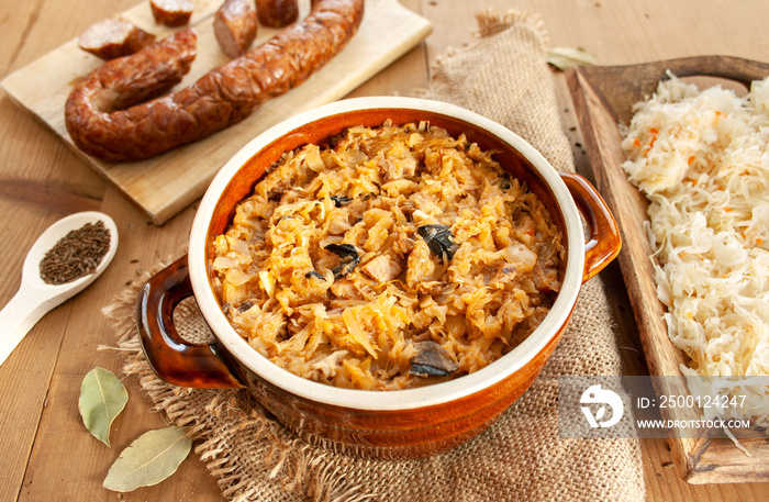 Bigos in stoneware dish, next to bigos ingredients: sausage, sauerkraut and cumin