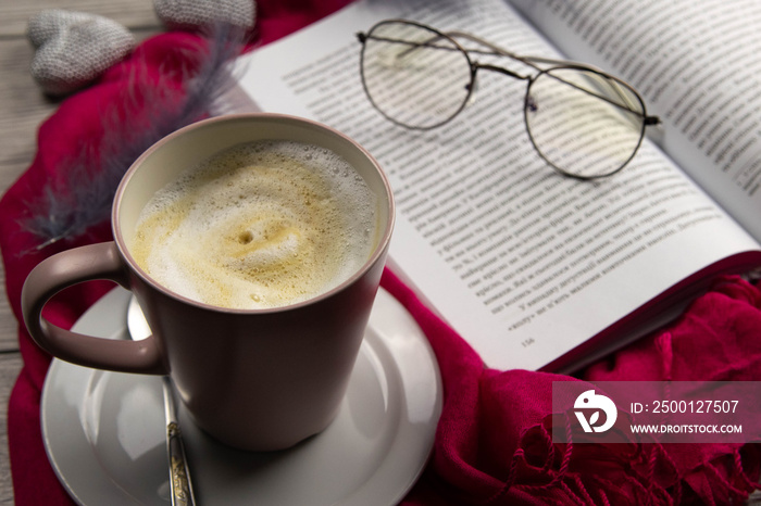 A mug of latte next to an open book on which there are glasses on the worm of a beautiful tablecloth