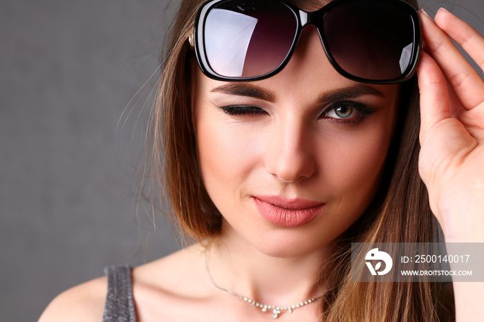 Beautiful young smiling brunette girl wearing sunglasses winking portrait over grey background. Modern sly coquette lady killer hunter plan concept