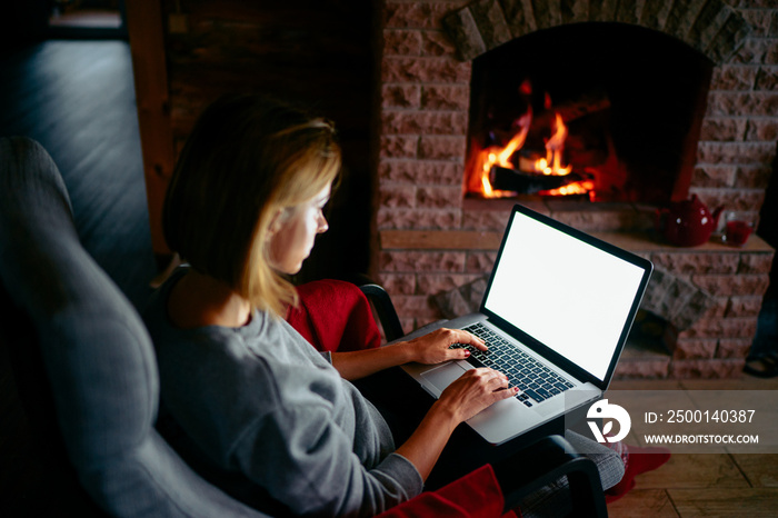 Cozy home. Pretty young woman working on laptop computer near the fireplace. Copy space on the screen.