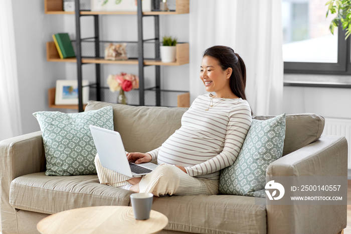 pregnancy, rest, people and expectation concept - happy smiling pregnant asian woman with laptop computer sitting on sofa at home