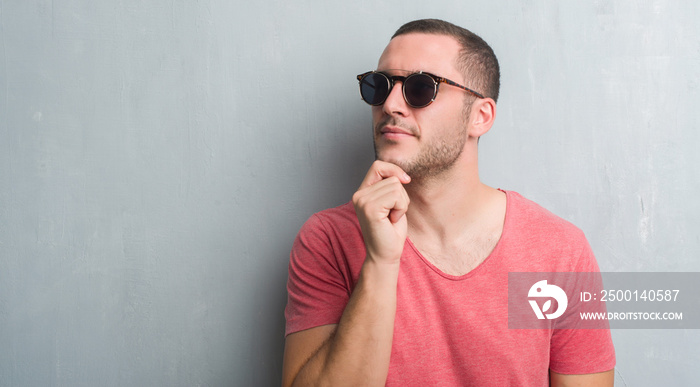 Young caucasian man over grey grunge wall wearing sunglasses serious face thinking about question, very confused idea