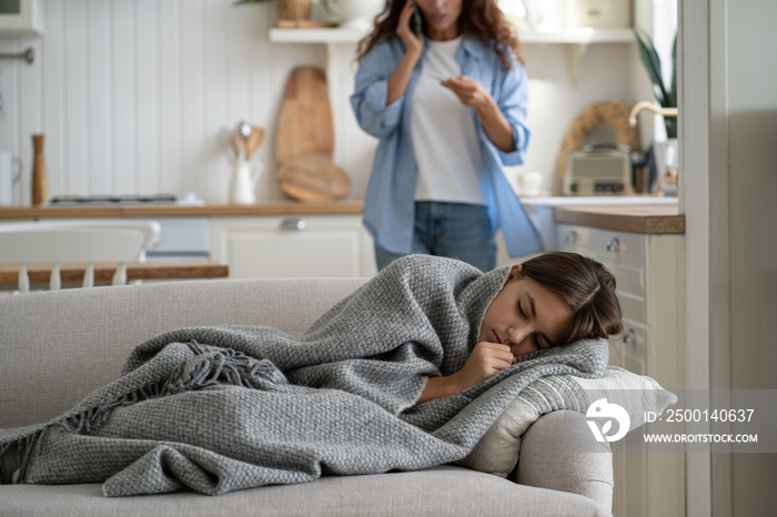 Sick child girl having fever and flu symptoms lying on sofa under blanket while her worried stressed mother talking to doctor by phone, calling to pediatrician, selective focus. Telehealth concept