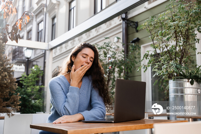 Sleeping girl with laptop is yawning outdoor on terrace in cafe. Tired young woman have a break at work and sleeping and relaxing at laptop on workplace due to overtime work.