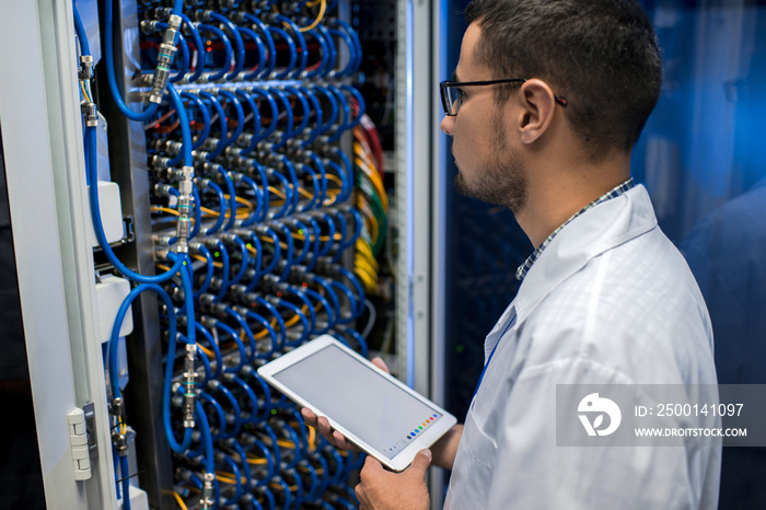 Portrait of young man holding digital tablet standing by supercomputer server cabinets in data center