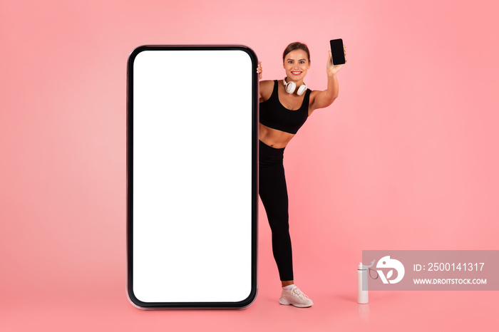 Check This. Excited Sporty Woman With Smartphone Peeking Behind Huge Blank Cellphone