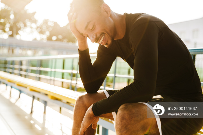 Image of smiling african american man sitting on bench at stadium