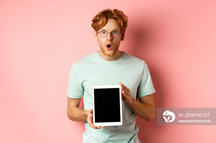 Excited young man with red hair and beard, checking out online promotion, showing digital tablet screen and staring at camera amazed, standing over pink background